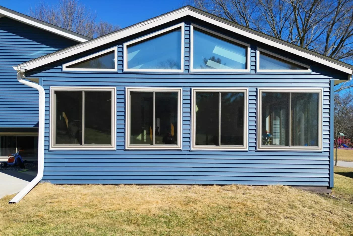 imgi_1_495091115_1242508717879867_2021652049905009721_n.webp Direct exterior view of a sunroom addition with blue horizontal siding, featuring four large sliding windows along the main wall and customized triangular transom windows fitted beneath the gabled roofline.
