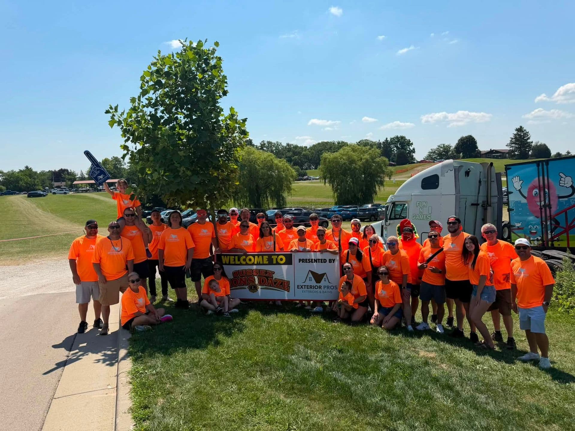 Group of volunteers wearing orange shirts holding a Welcome to Sunken In Daze banner at an outdoor event in a park with a trailer and recreational vehicles visible in the background