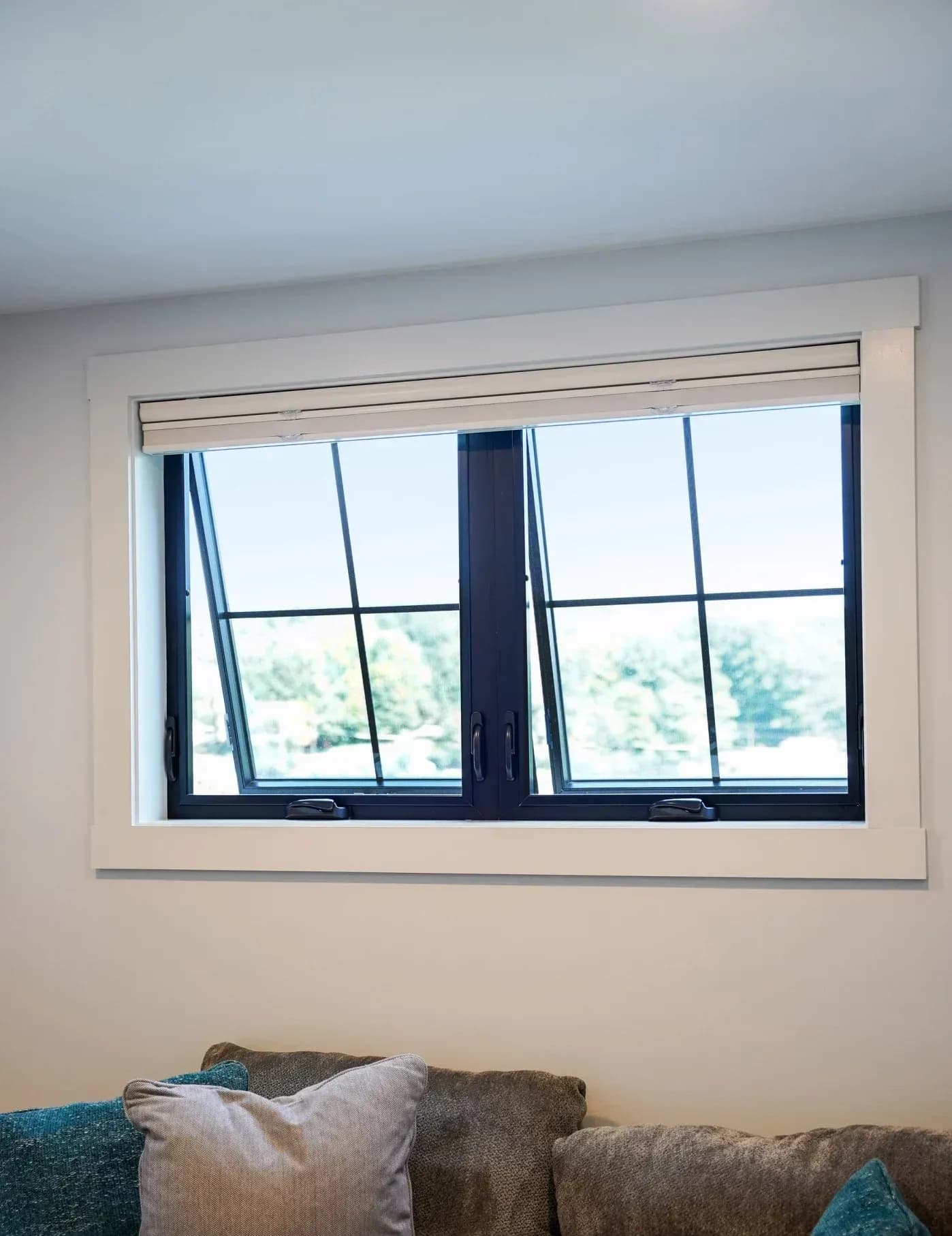 Interior view of two side-by-side dark-framed awning windows with simple window grids, crank handles, and crisp white trim, shown slightly pushed open at the bottom above a sofa.