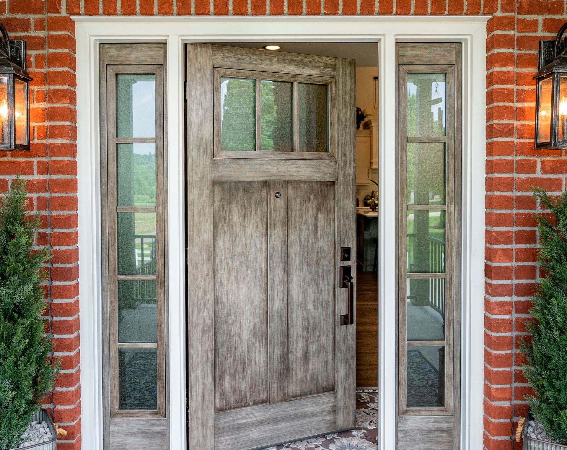 Craftsman-style front door with transom window and sidelights, gray wood finish, set in red brick exterior with white trim and lantern sconces