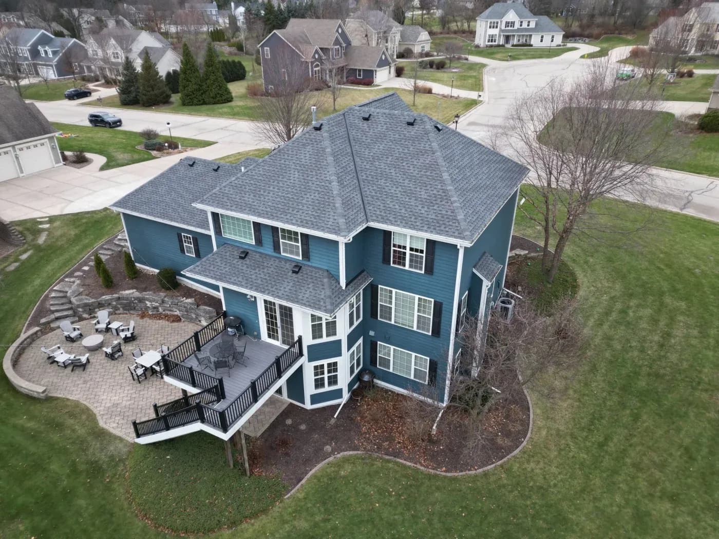 Aerial view of a blue two-story colonial home with gray roof, featuring a wraparound patio, multiple windows, and landscaped yard in suburban neighborhood