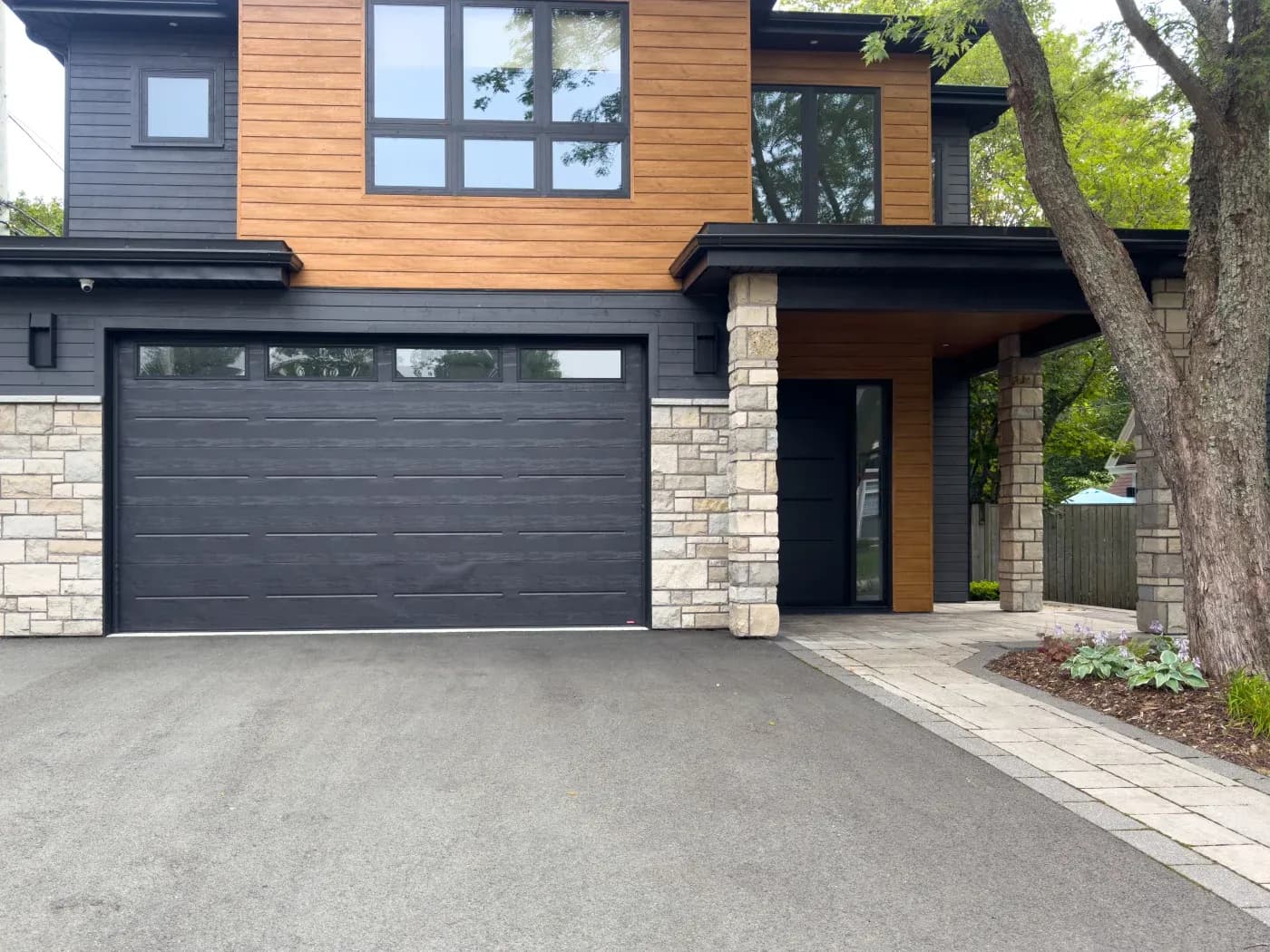 Exterior view of a modern two-story home featuring black vertical/horizontal siding paired with warm wood-look siding panels, light-colored faux stone accents around the garage base and porch pillars, a dark garage door, and a matching front door.