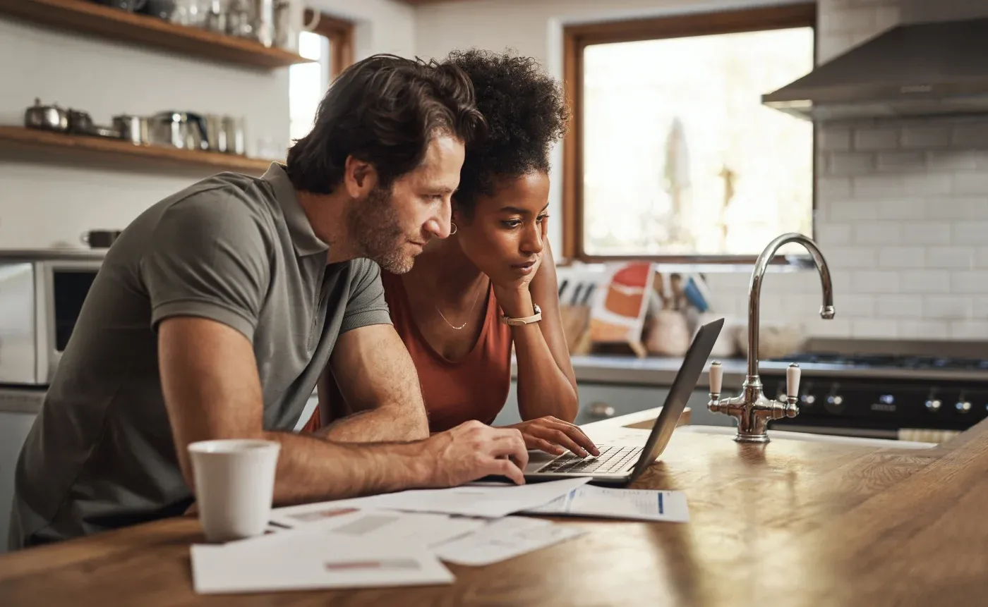 Interior view of a couple sitting together at a wooden kitchen island, attentively leaning over a laptop screen with financial documents spread out in front of them.