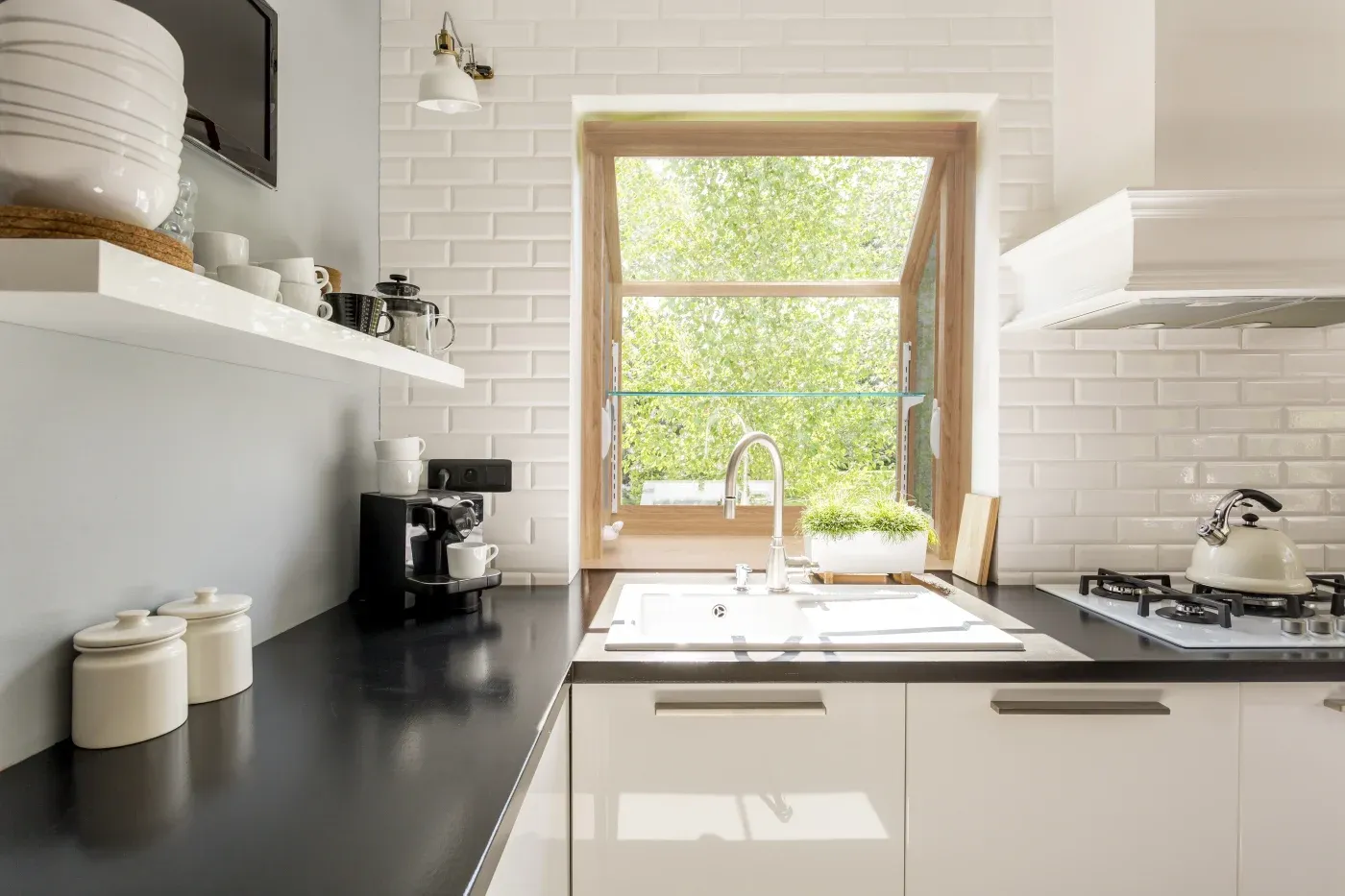 Interior view of a modern kitchen featuring a light wood-framed garden window above a white sink and gooseneck faucet, set into a white subway tile backsplash with black countertops and white cabinetry.