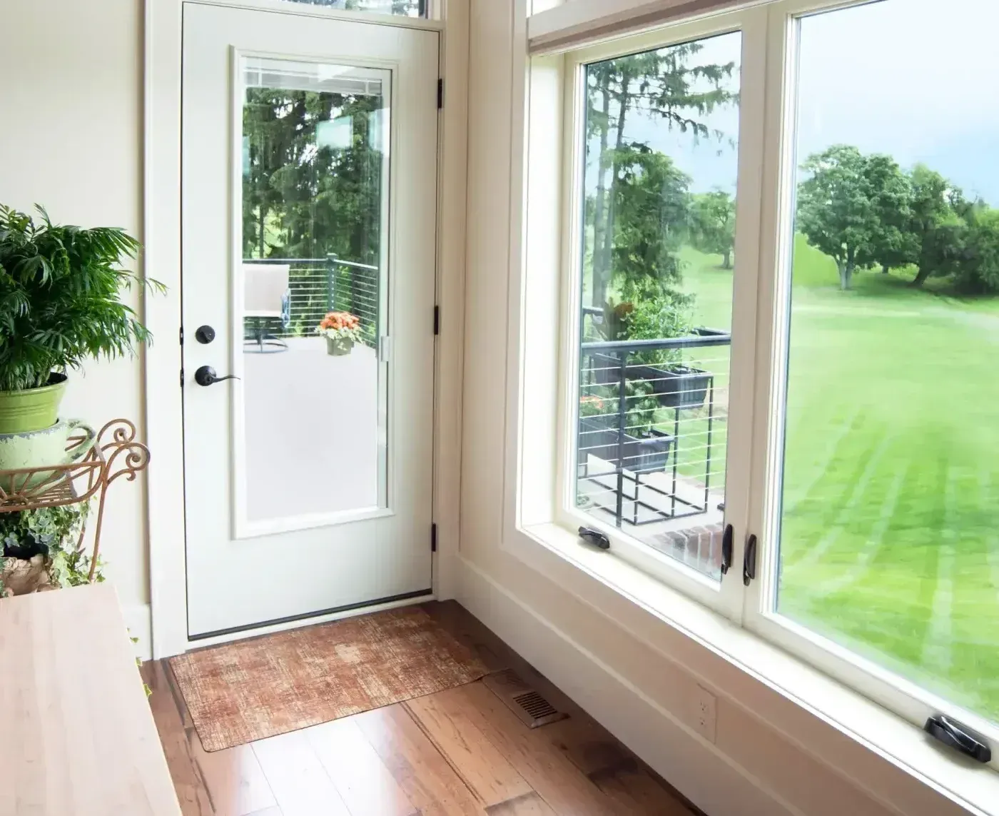 Interior view of a sunlit entryway featuring white-framed casement windows with crank handles, looking out over a green lawn, adjacent to a white patio door with glass insert leading to a deck with cable railing.