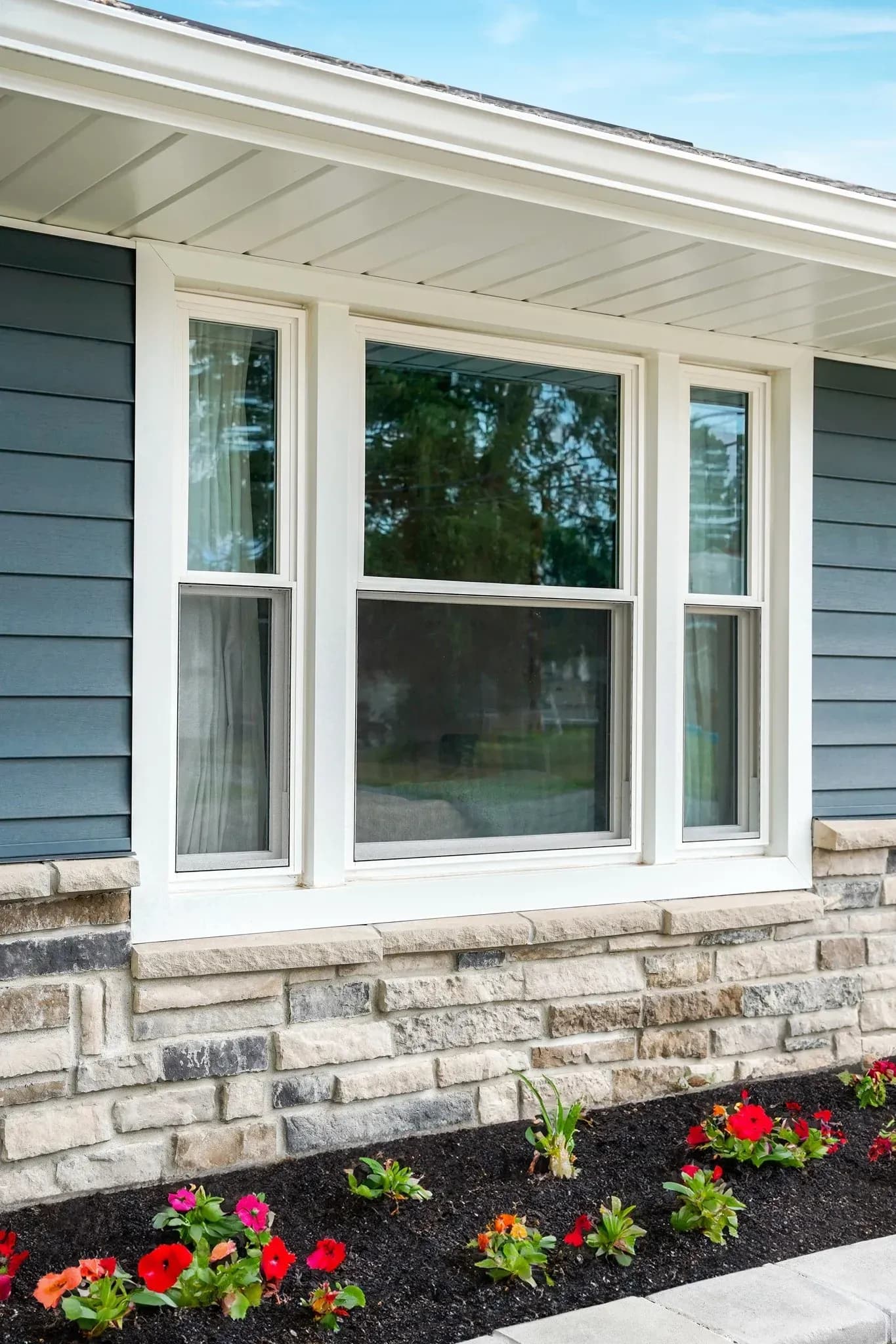 Exterior view of a modern home featuring dark blue lap siding above a stacked stone ledge, showcasing a set of white vinyl double-hung windows flanked by narrower side windows, set over a flower bed with mulch and colorful annuals.