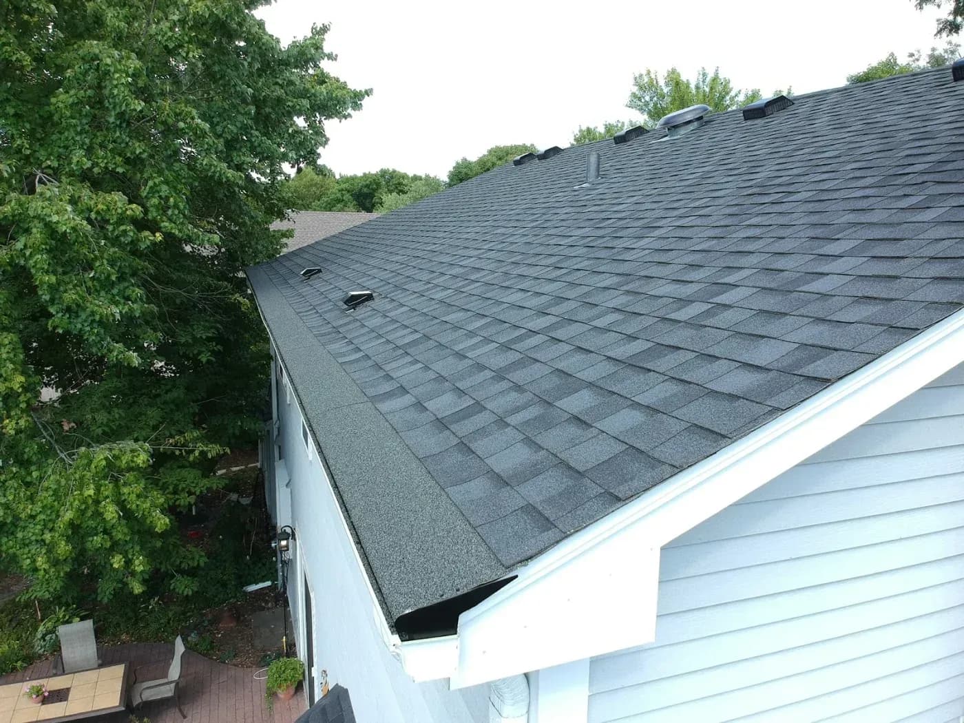 High-angle view of a grey shingled home roof line featuring installed dark metal gutter covers and roof vents, situated above a patio with surrounding leafy trees.