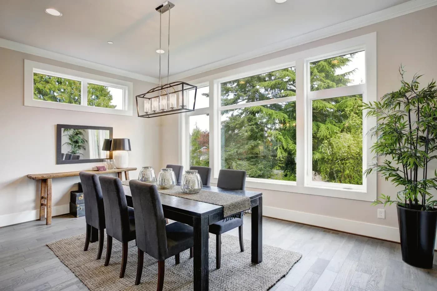 Interior view of an elegant dining room featuring large floor-to-ceiling picture windows with white trim looking out onto lush green trees, a modern dining set with dark upholstered chairs, a rectangular pendant light, and a high horizontal transom window on the side wall.