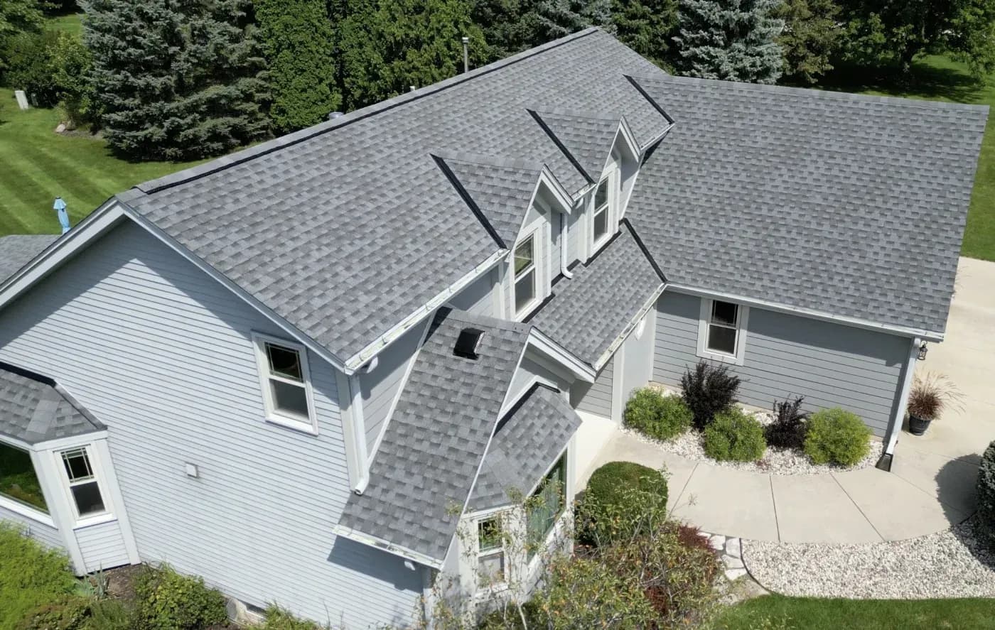 High-angle aerial view of a light grey siding home featuring a newly installed multi-gabled grey architectural shingle roof, dormer windows, and landscaped walkway.