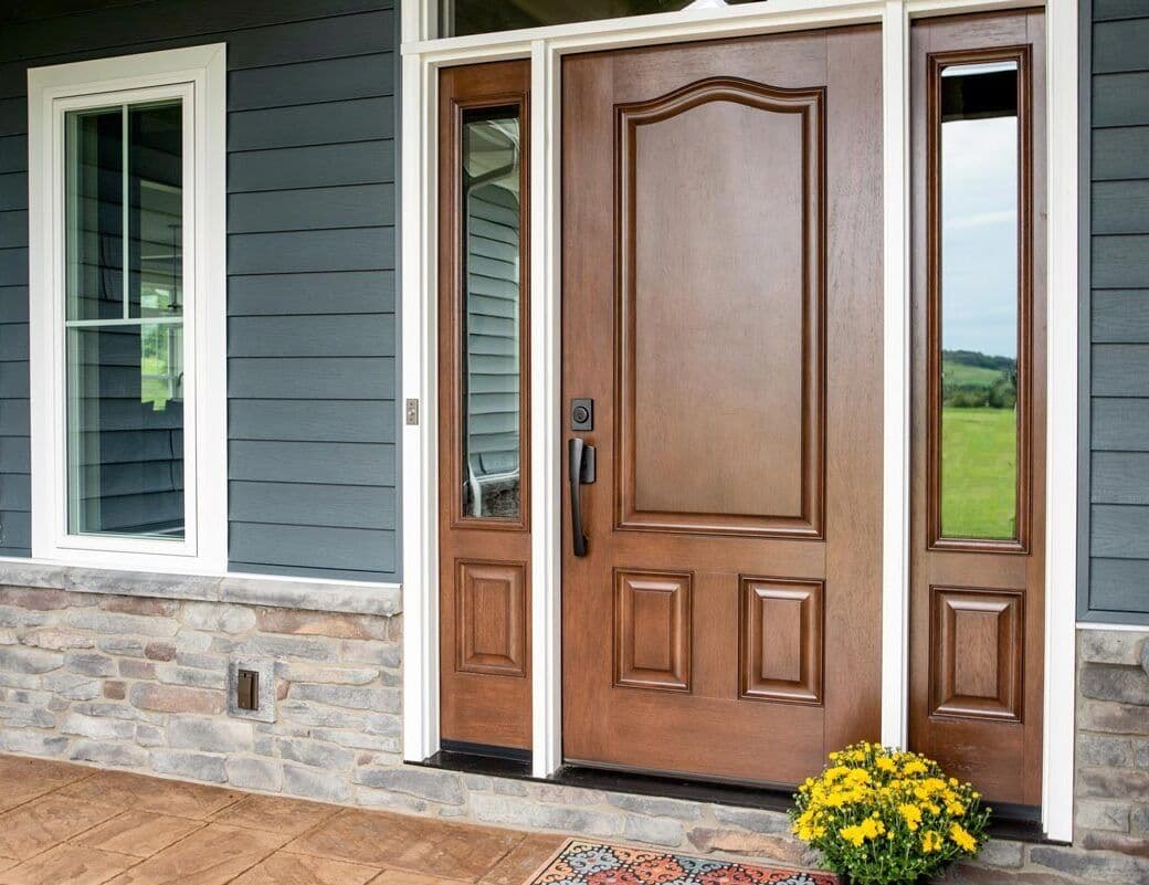 Exterior view of a home's front entrance featuring a rich hazelnut wood-grained entry door with raised paneling and matching full-length glass sidelights, framed by crisp white trim against dark slate-grey horizontal siding and a stacked stone base wall.