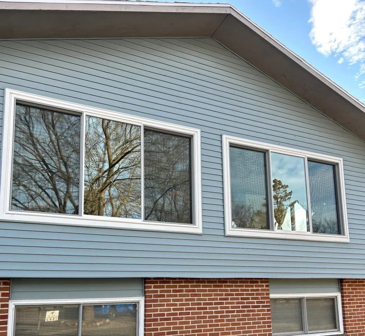 Exterior view of a home's upper story with blue horizontal siding featuring two large 3-lite sliding windows with white frames, positioned above a lower level with red brick veneer and sliding basement windows.
