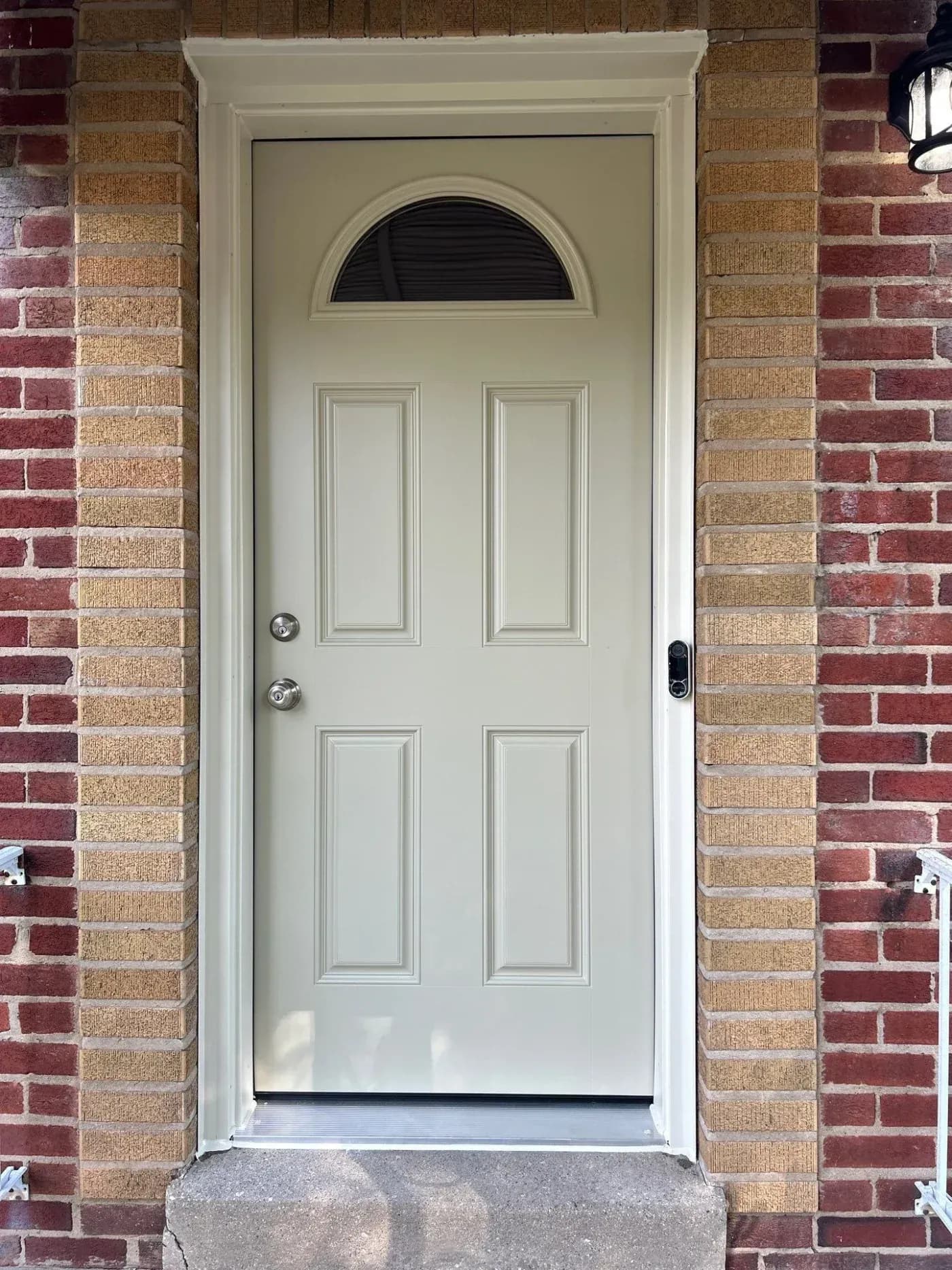 Light beige front entry door with raised panels, a half-moon window, silver hardware, and white trim set in a brick exterior.