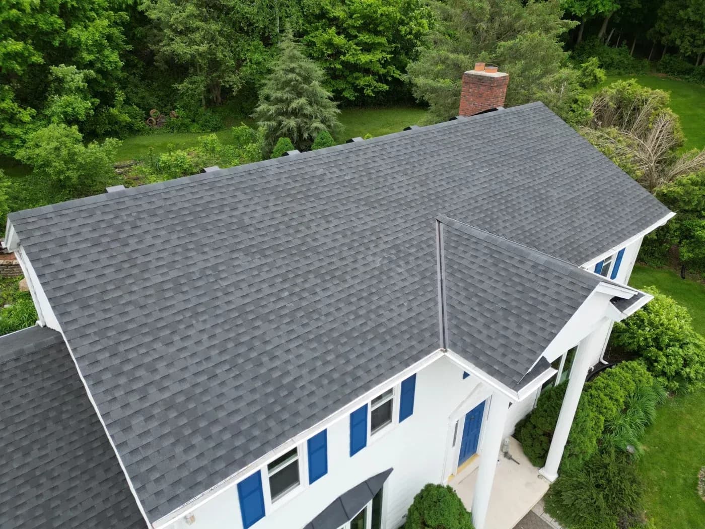 Aerial view of a white two-story home with a newly installed gray asphalt shingle roof, brick chimney, blue shutters, and surrounding trees.