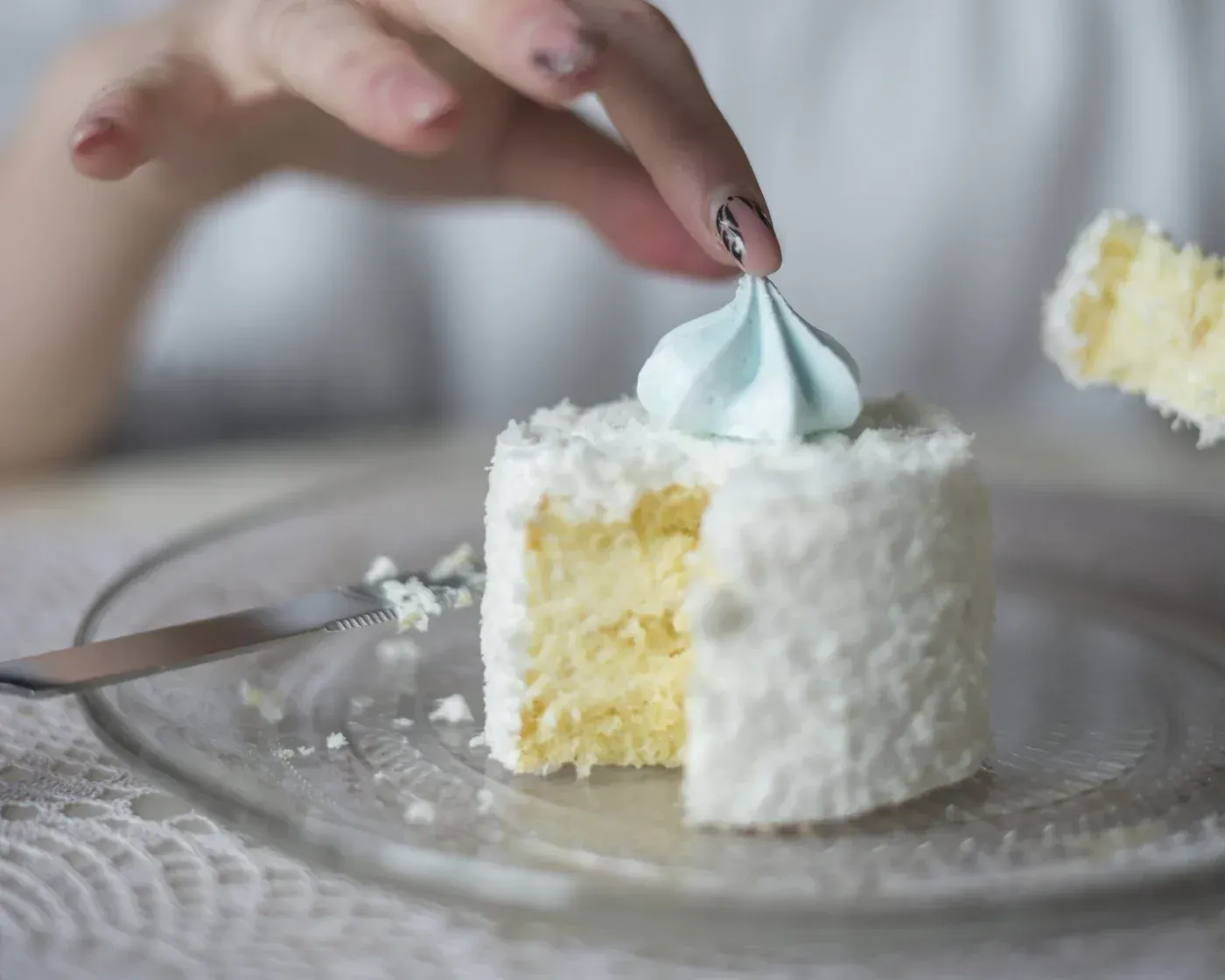 Close-up of a hand placing a decorative meringue on a slice of white frosted cake.