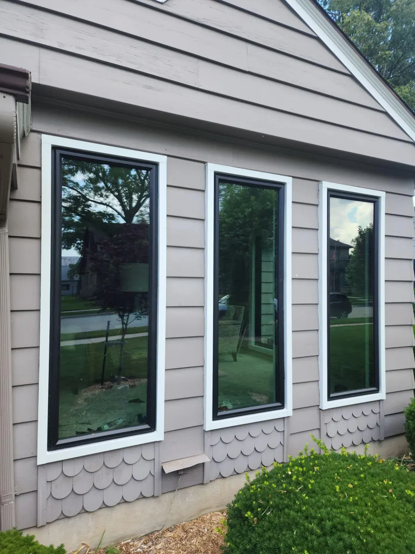 Three tall black-framed replacement windows with white trim installed on a beige-sided home exterior.