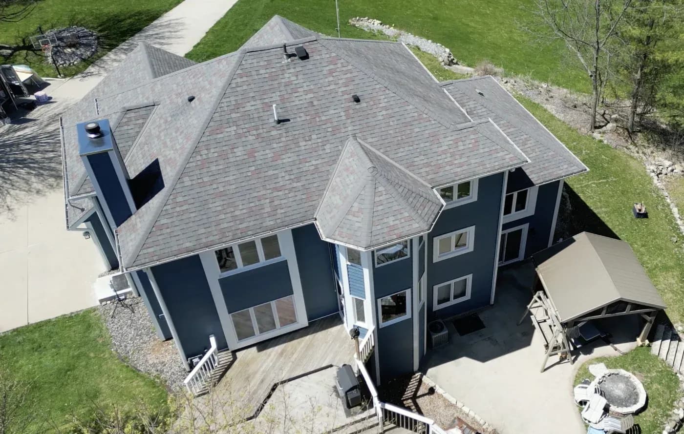 Aerial view of a large blue two-story home with a multi-level asphalt shingle roof, rear deck, patio, and gazebo.