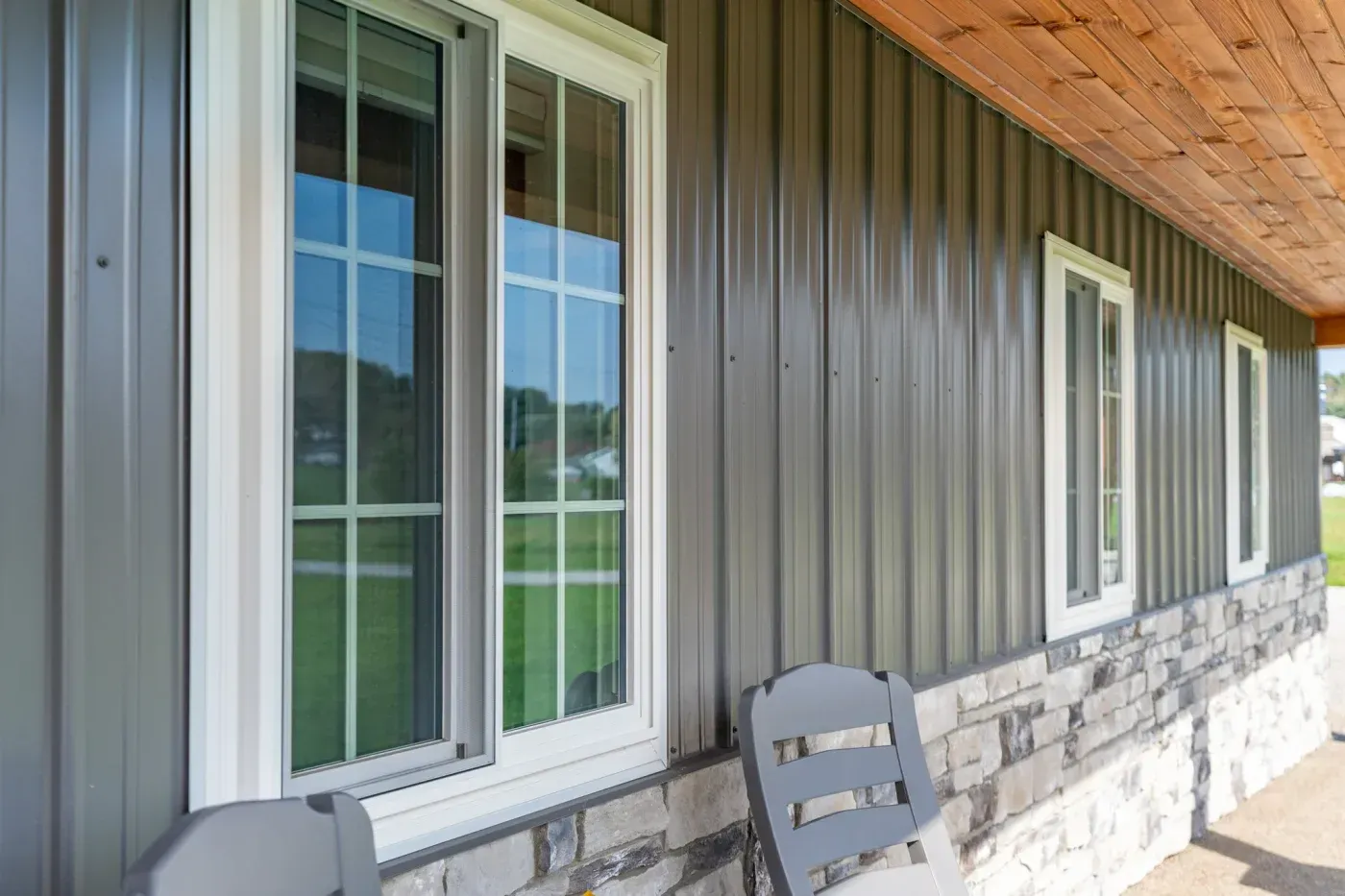 Exterior view of a home with dark vertical metal siding, white-trimmed replacement windows, stone veneer, and a wood-paneled porch ceiling.