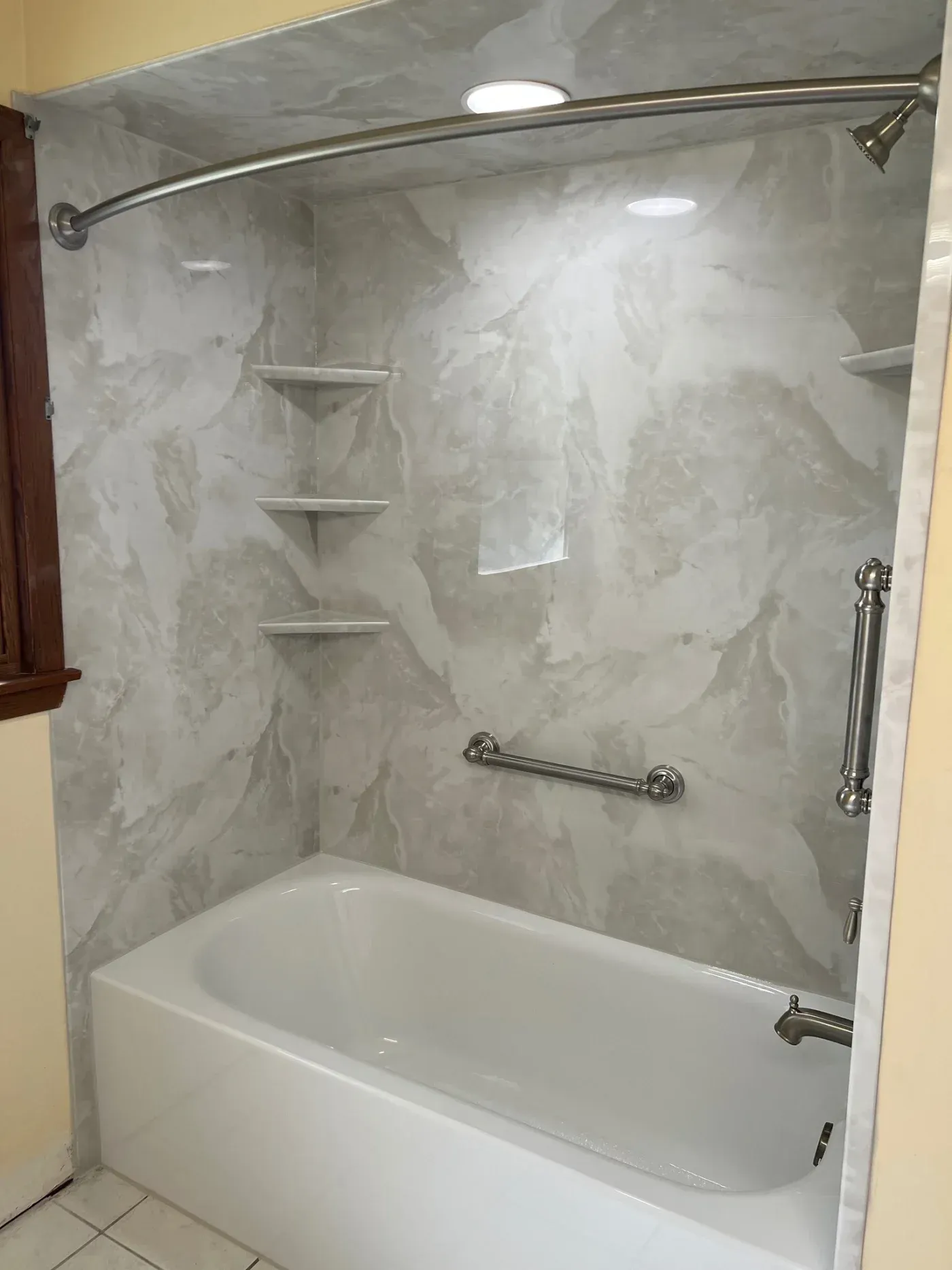 White bathtub and shower surround with marble-look wall panels, built-in corner shelves, grab bars, and brushed metal fixtures.