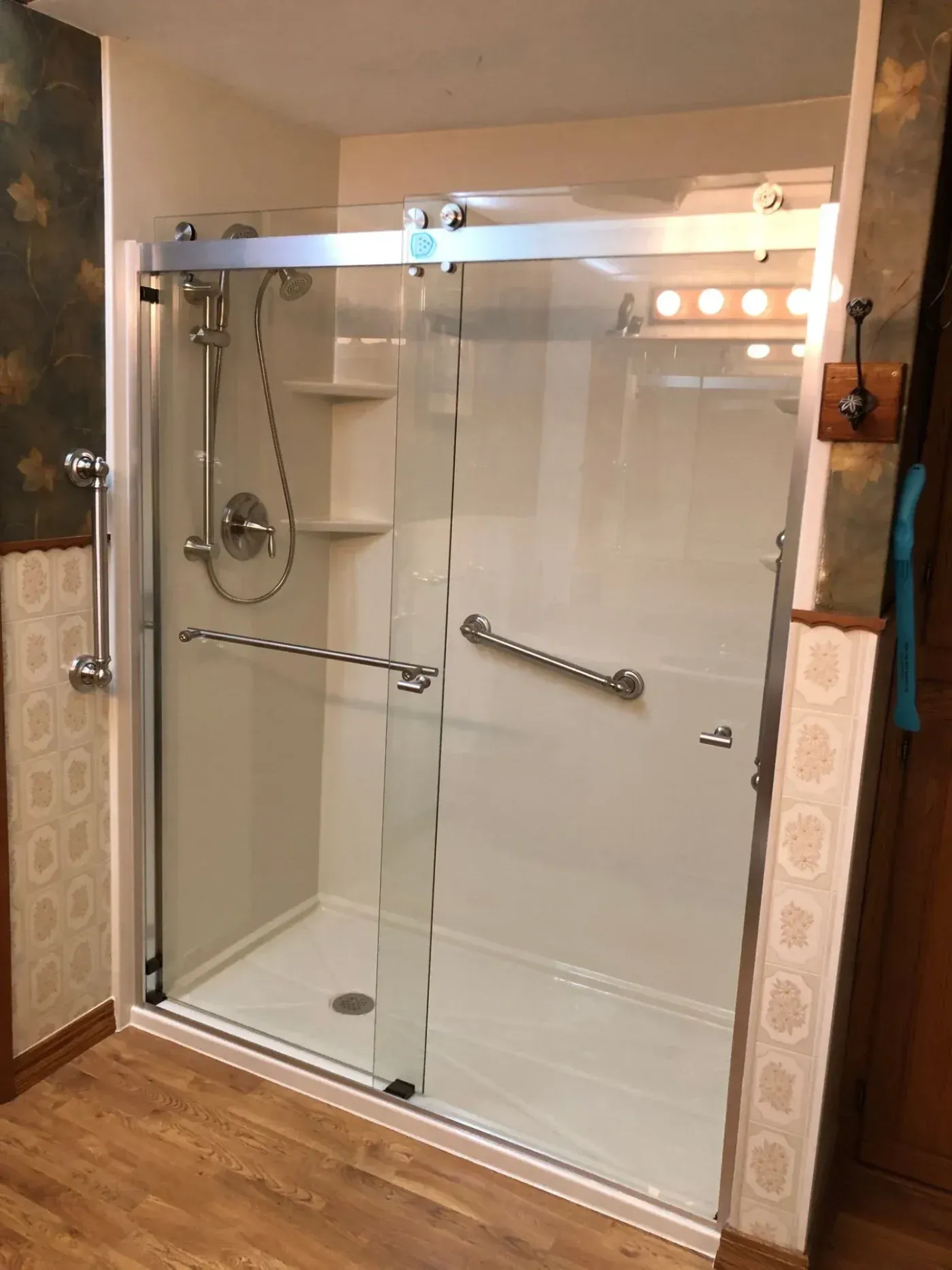 Tub-to-shower conversion with white wall panels, sliding glass doors, handheld showerhead, built-in corner shelves, and safety grab bars in Allenton, Wisconsin.
