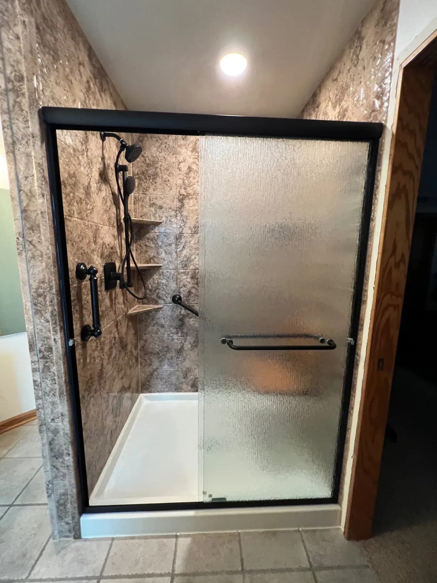 Walk-in shower replacement with stone-look wall panels, textured glass sliding door, black fixtures, grab bars, and built-in corner shelves in Ashippun, Wisconsin.