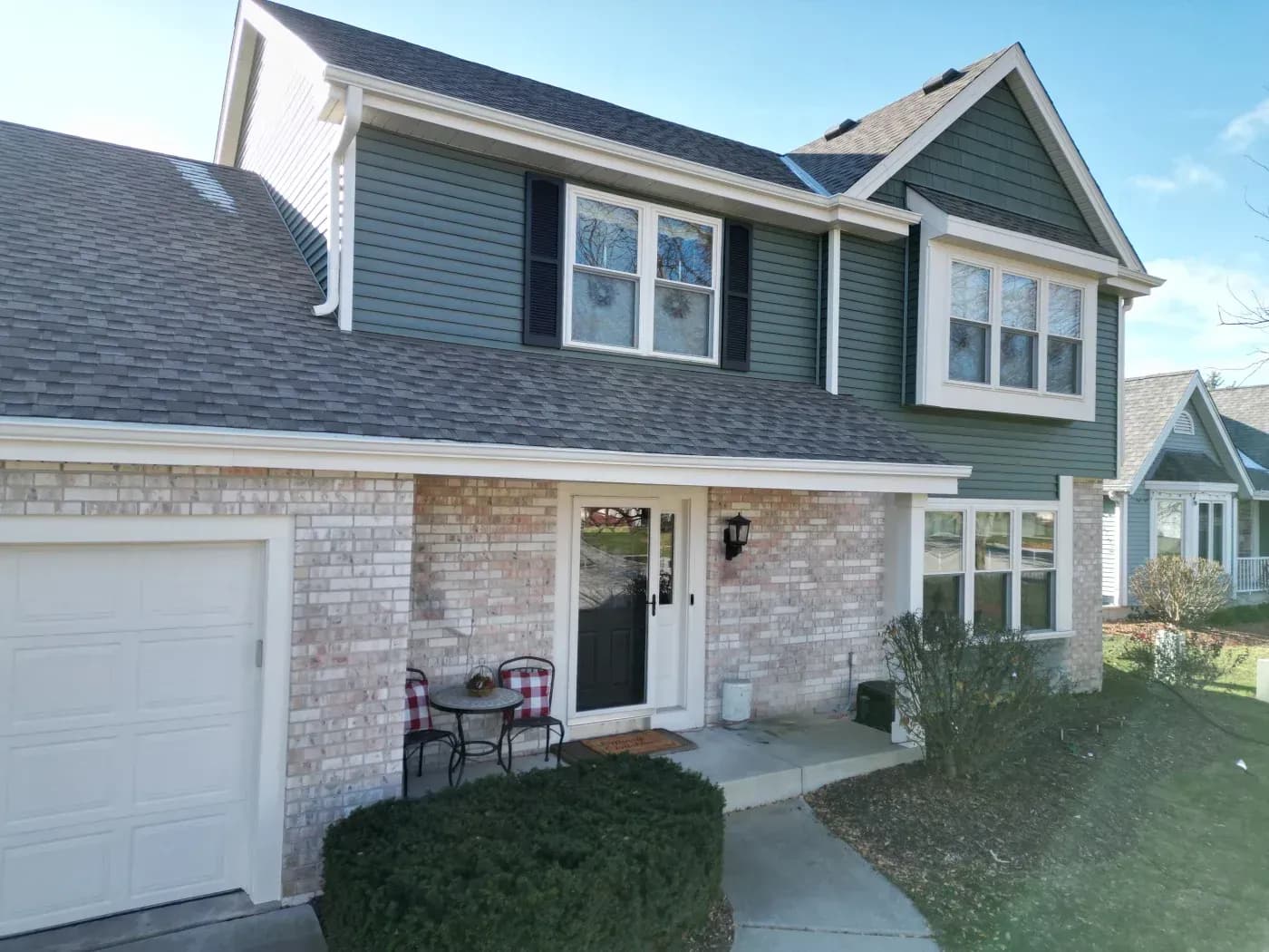 Two-story home with green vinyl siding, white trim, brick exterior, replacement windows, asphalt shingle roofing, and gutters in Pewaukee, Wisconsin.