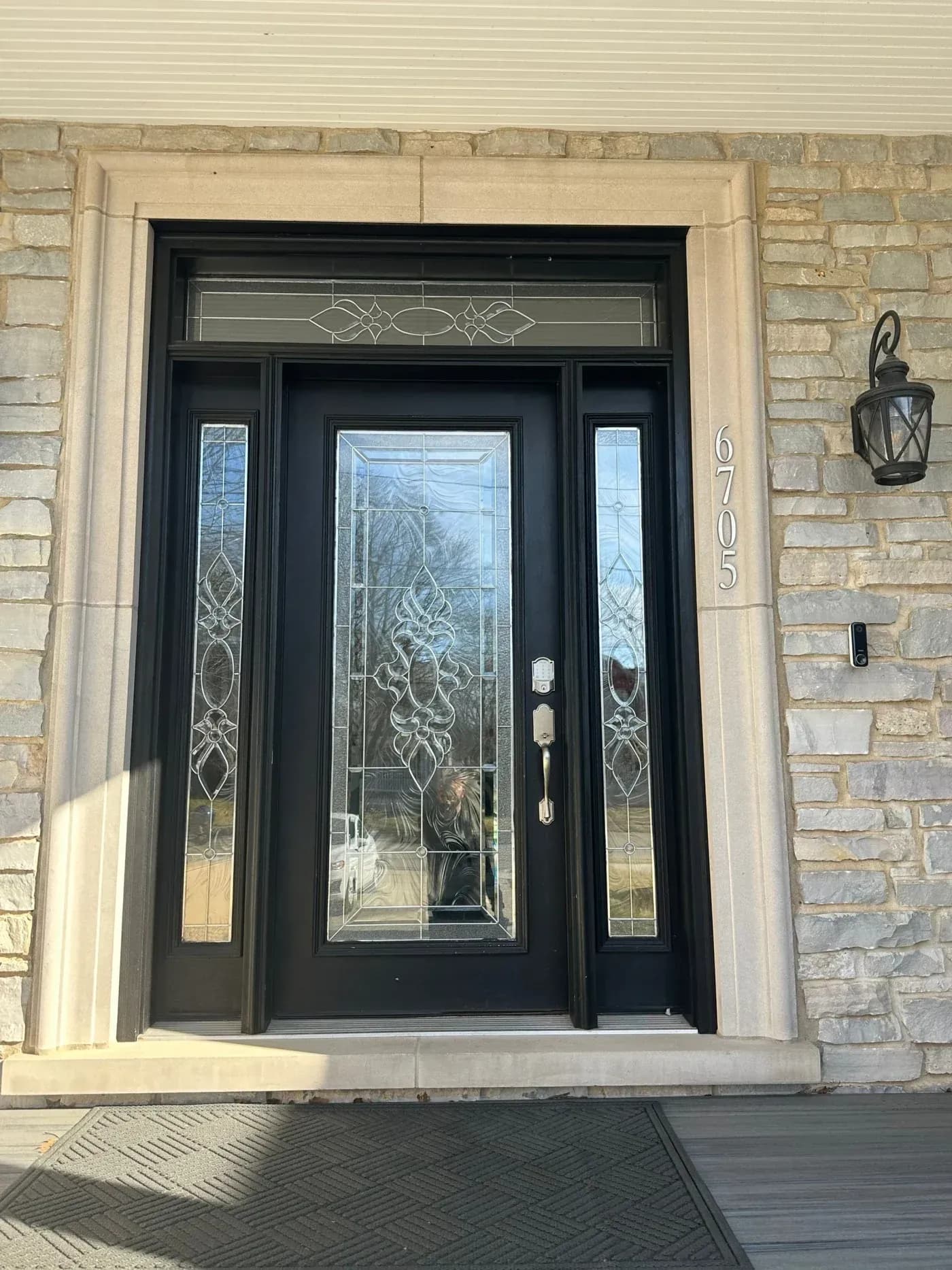 Black front entry door with decorative glass panel, matching sidelights and transom, set in a stone exterior with beige trim.