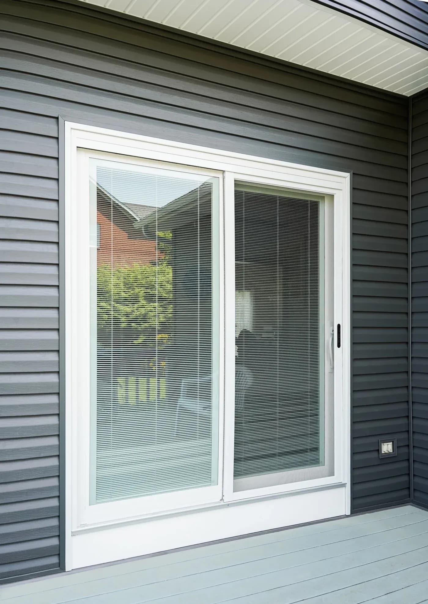 Exterior view of a white vinyl sliding patio glass door set into dark grey lap siding, featuring built-in mini blinds enclosed between the glass panes.