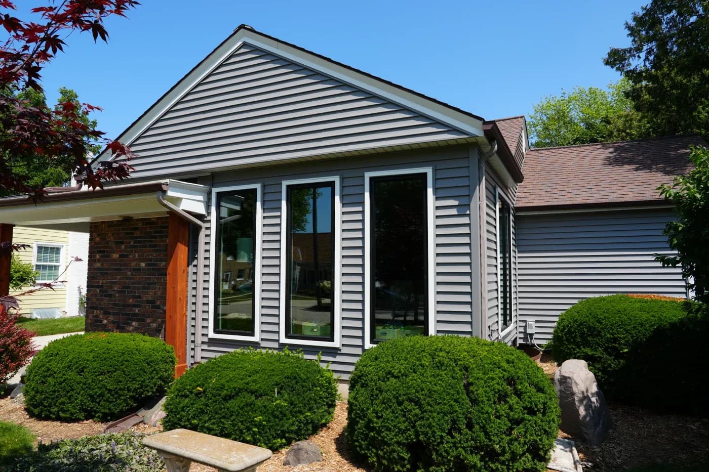 Exterior view of a home featuring grey vinyl horizontal lap siding, a brick accent wall under a covered porch entry, three tall floor-to-ceiling windows with black frames and white exterior trim, and manicured green shrubbery.