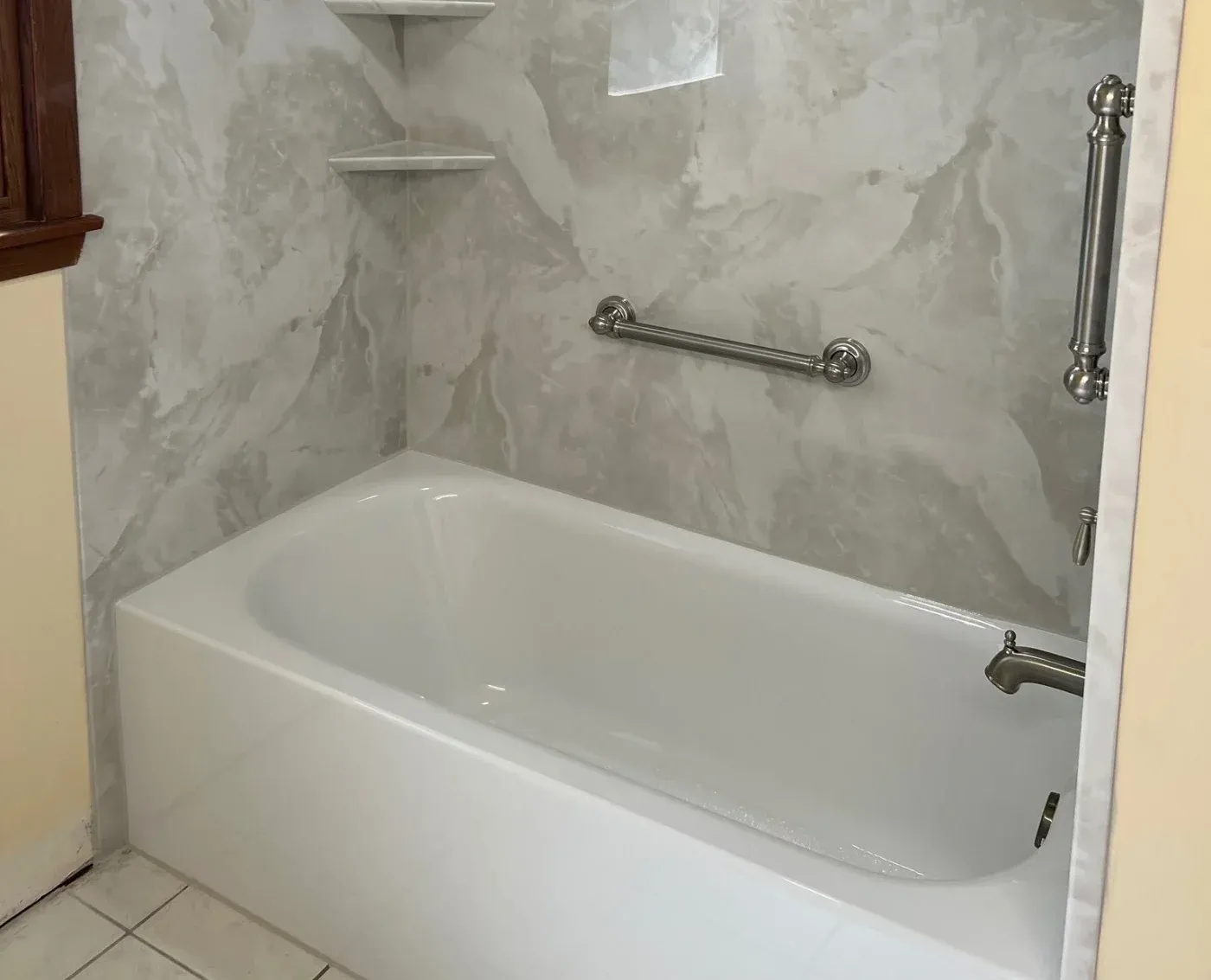 Interior view of a remodeled alcove bathtub featuring a crisp white tub basin, faux marble wall panels, brushed metal fixtures including a grab bar and tub faucet, and corner storage shelves.