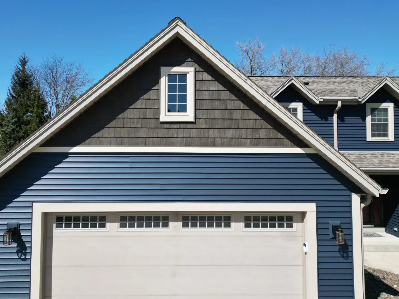 Exterior view of a home's gable roof section above a garage, featuring deep blue horizontal vinyl siding paired with a dark grey shake-style siding accent peak, highlighted by crisp white trim around a small accent window and garage door frame.