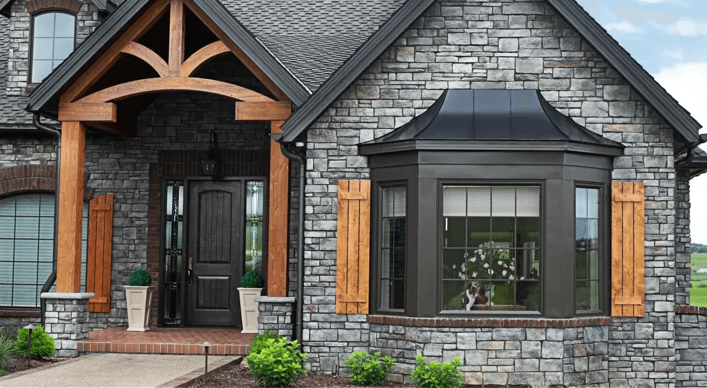 Modern stone home exterior featuring arched wooden entry portico with black door, bay window with black metal roof, cedar shutters, and landscaped front foundation